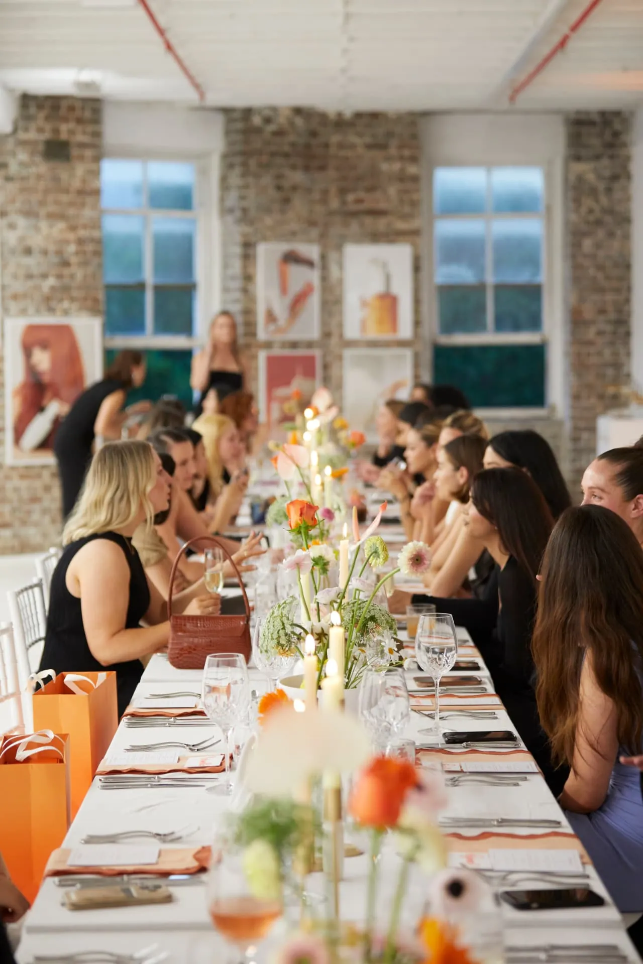 Guests seated along a long candlelit table at a Sydney public event, with floral centrepieces and orange gift bags.