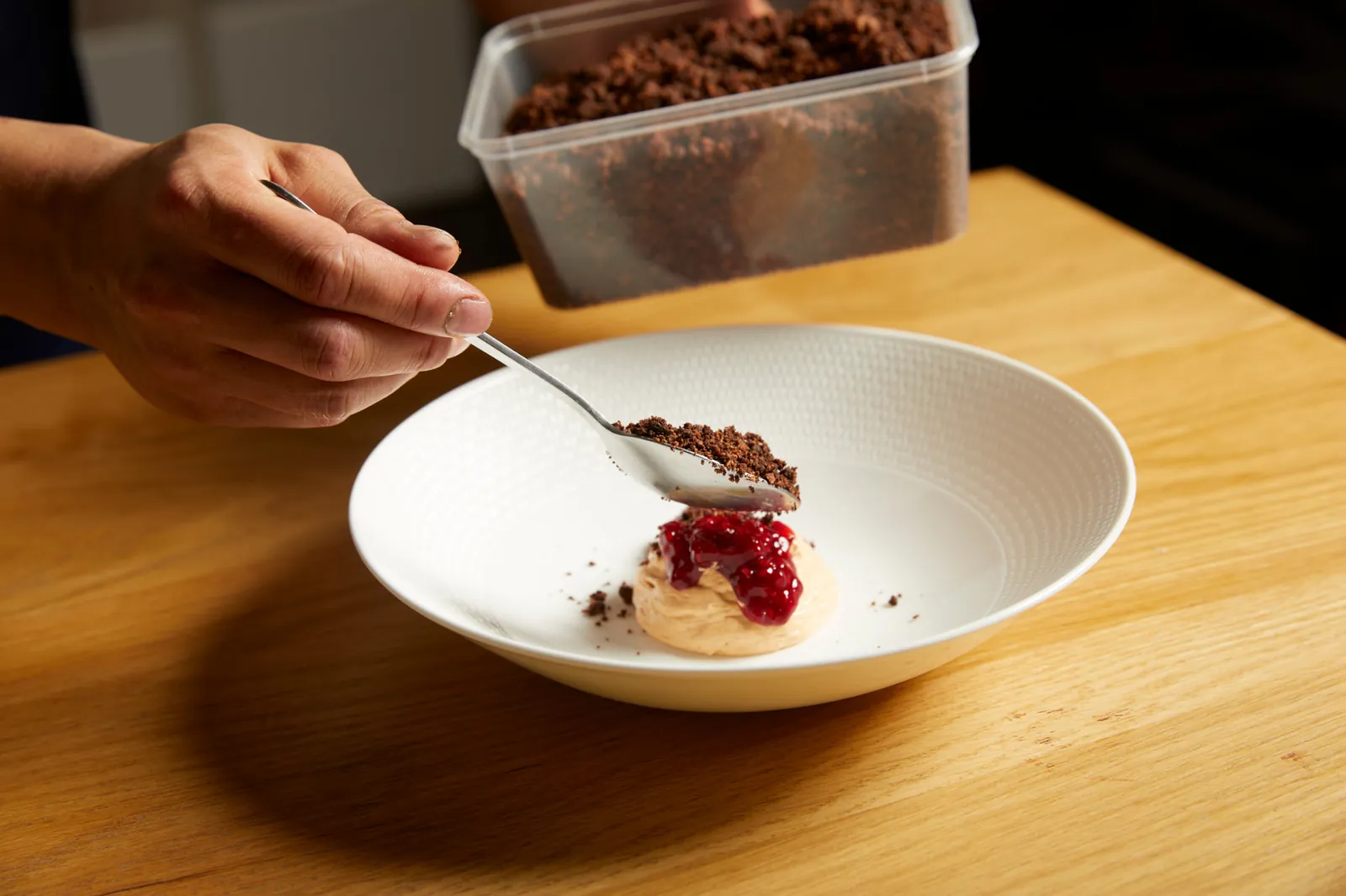 Chef's hand sprinkling chocolate crumble onto a meringue with berry compote in a white bowl.