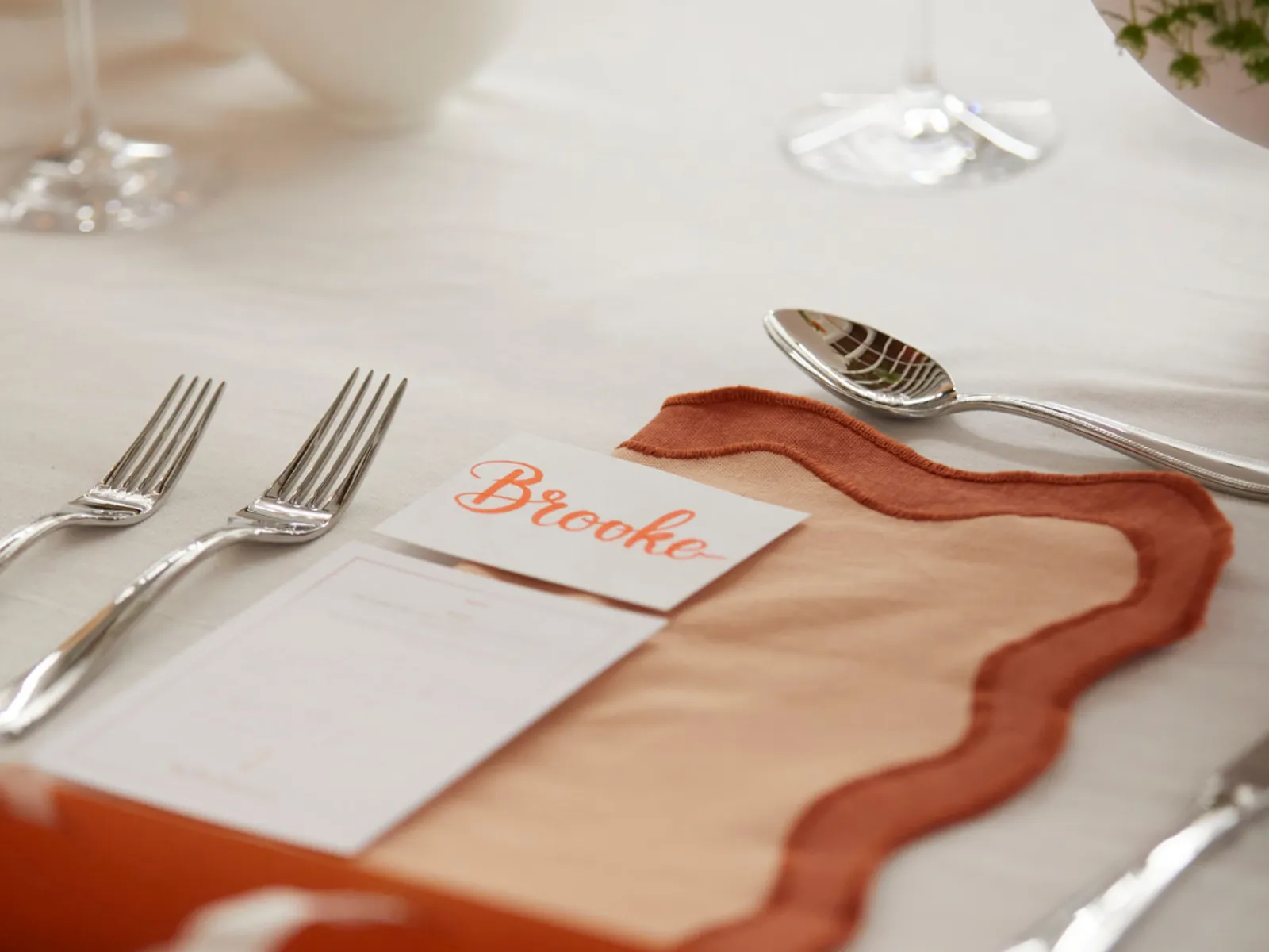 An elegant executive dinner place setting with a handwritten name card, crystal glassware and terracotta linen on a white tablecloth.