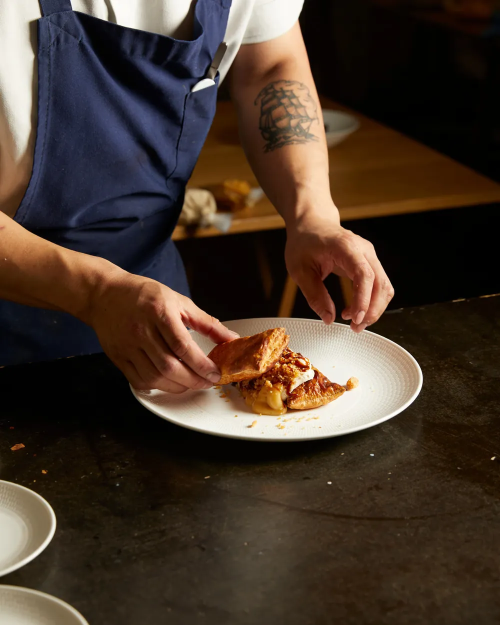 A Kreative Catering chef carefully finishing a flaky pastry dish on a white plate, sleeves rolled up in a navy apron.