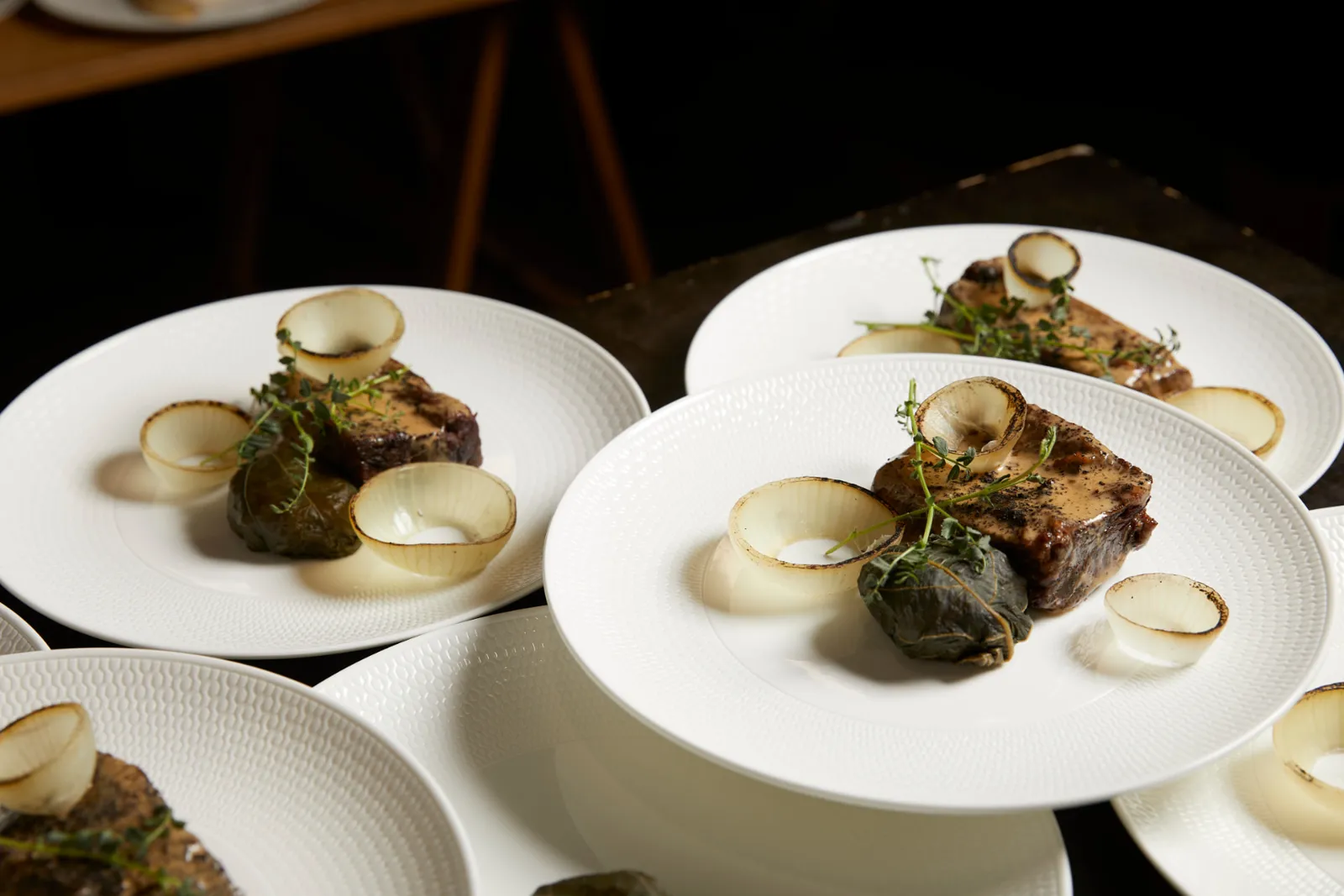 Plated beef with thyme, charred onion petals and vine-leaf parcel on white dinner plates.