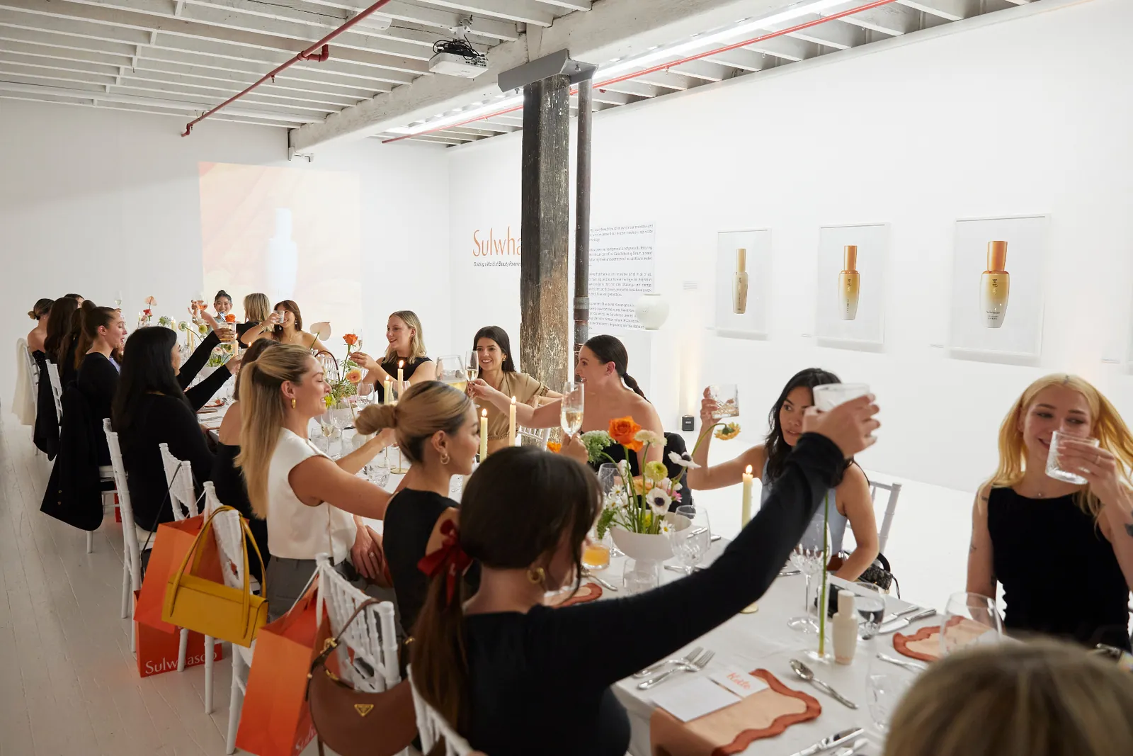 Guests raising glasses around a long dressed table in a bright white gallery venue with tall candles and floral centrepieces.